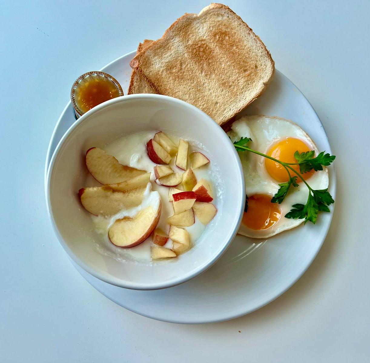 A photograph of a breakfast plate at InterCube hostel in Dubai, featuring two sunny-side-up eggs garnished with parsley, a bowl of yogurt topped with sliced apples, and two pieces of toasted bread with a small jar of jam