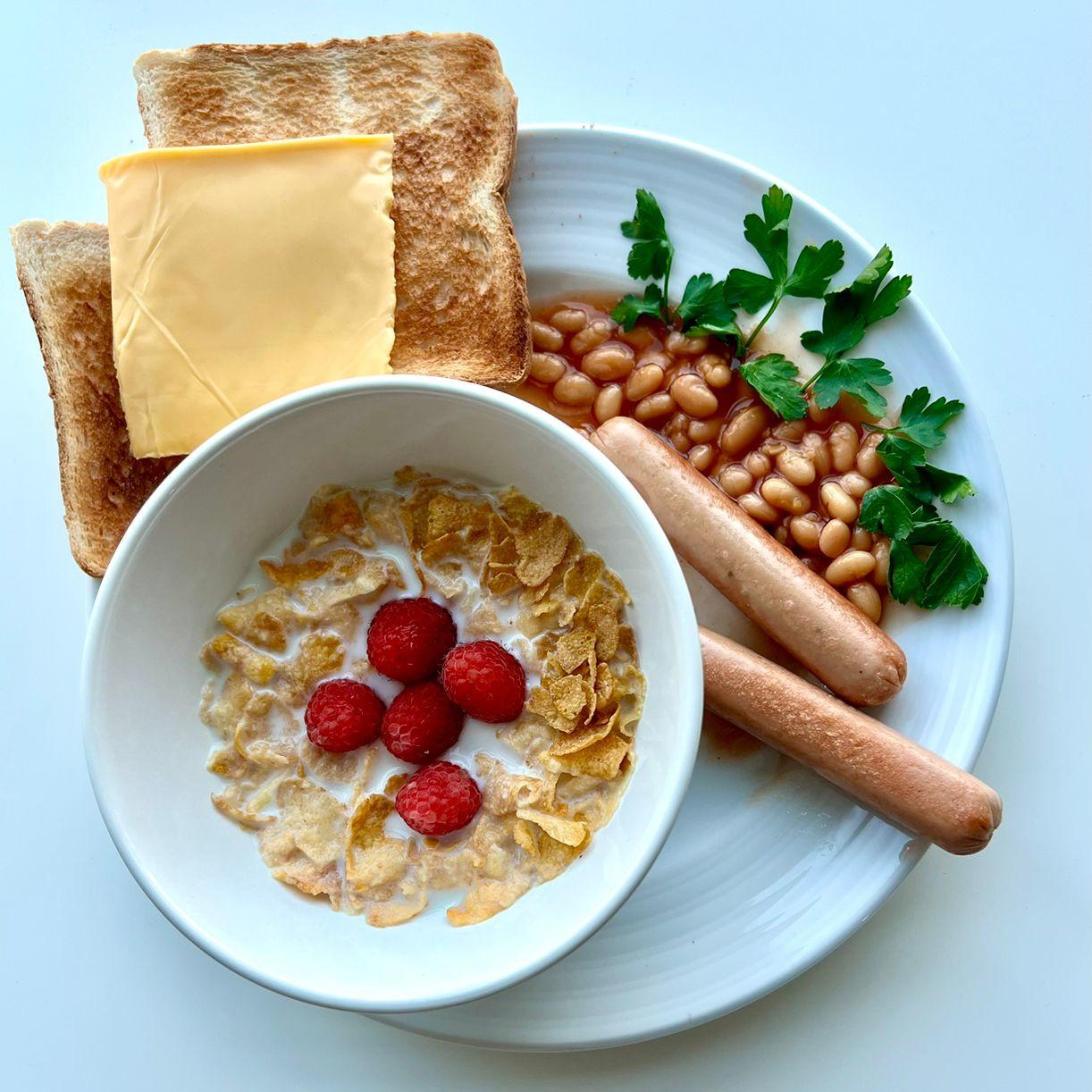 A photograph of a breakfast plate at InterCube hostel in Dubai, featuring two pieces of toasted bread with a slice of cheese, a bowl of cereal topped with raspberries, two sausages, baked beans garnished with parsley